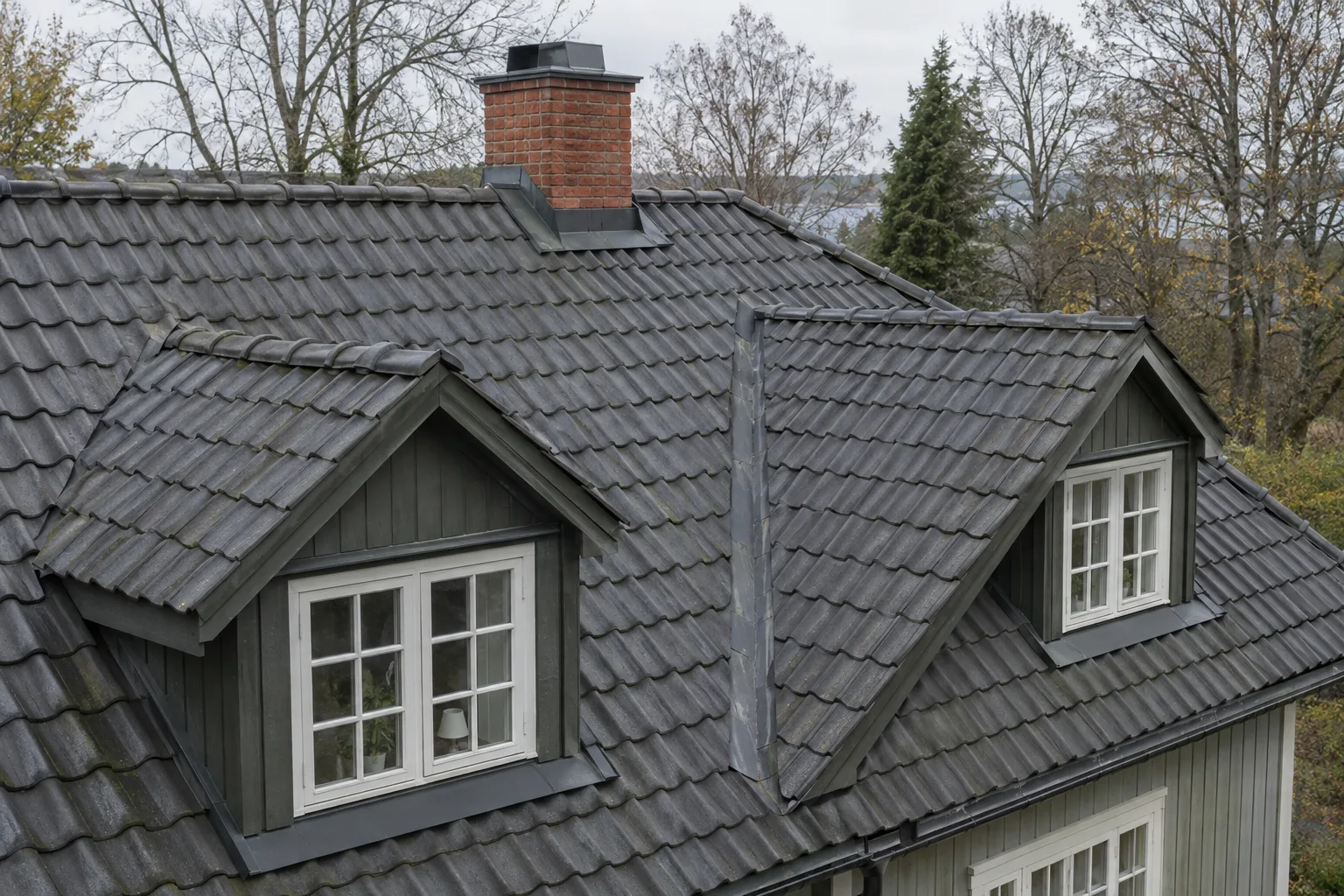Mörkt tegeltak på en villa med två takkupor, en vinkelränna och murad skorsten.